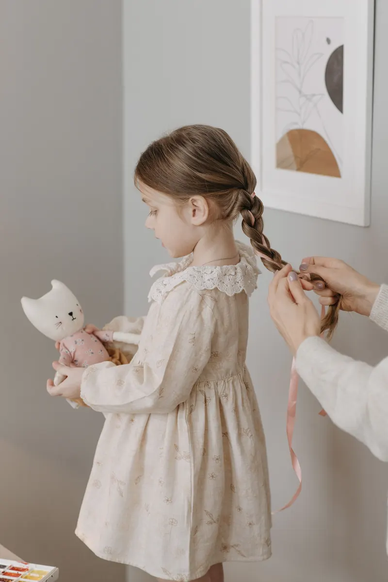 A young girl having her hair gently braided indoors, wearing a white dress